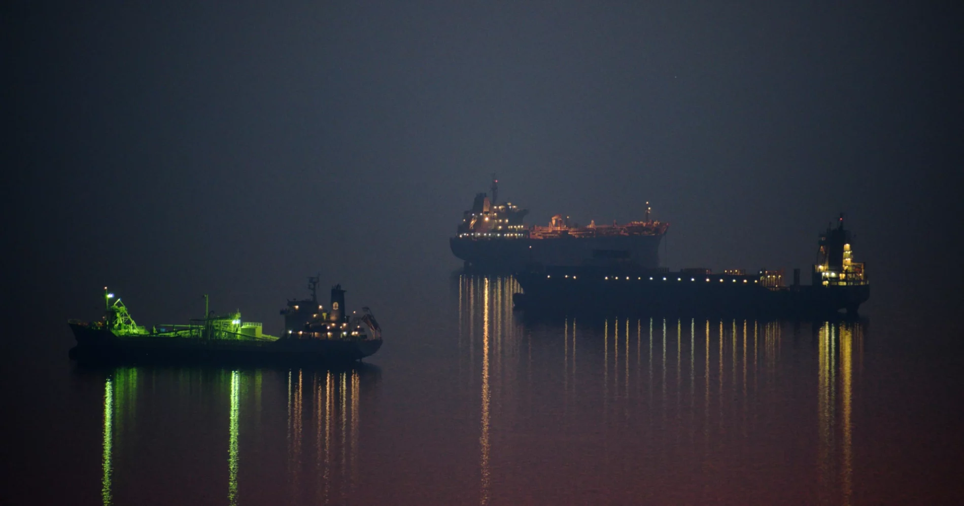 Three illuminated cargo ships at sea at night, with lights reflecting on the water under a foggy sky.