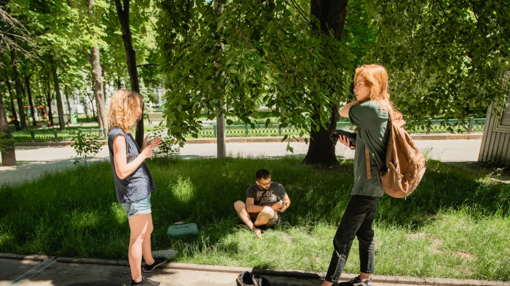 Three people are outside in a park - the group is positioned in a triangular arrangement. Two women stand and talk on a surfaced pathway in profile to the camera. Next to them, facing towards the camera, a man is sitting corss-legged on a grassy area, shaded by some trees.