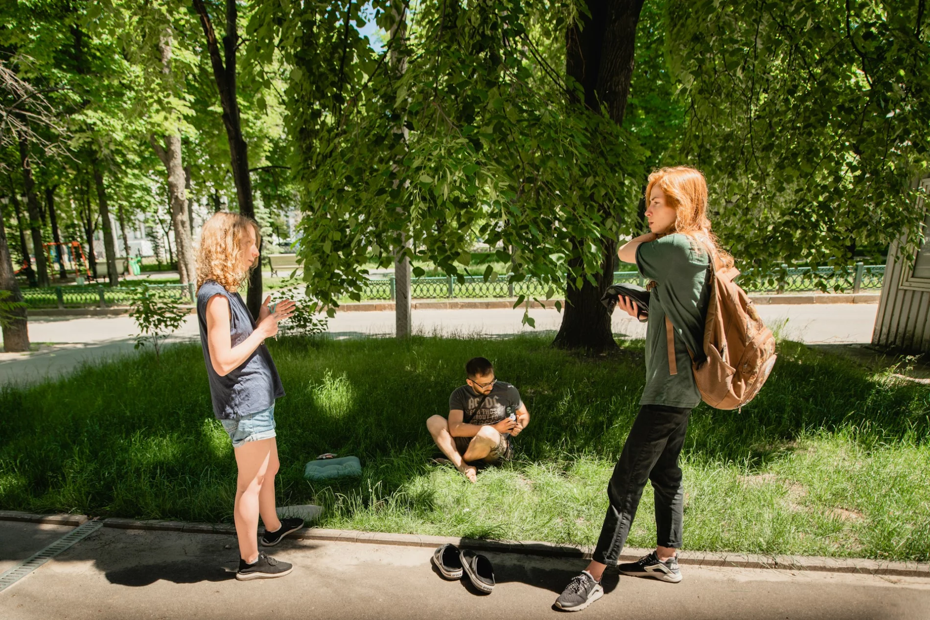Three people are outside in a park - the group is positioned in a triangular arrangement. Two women stand and talk on a surfaced pathway in profile to the camera. Next to them, facing towards the camera, a man is sitting corss-legged on a grassy area, shaded by some trees.