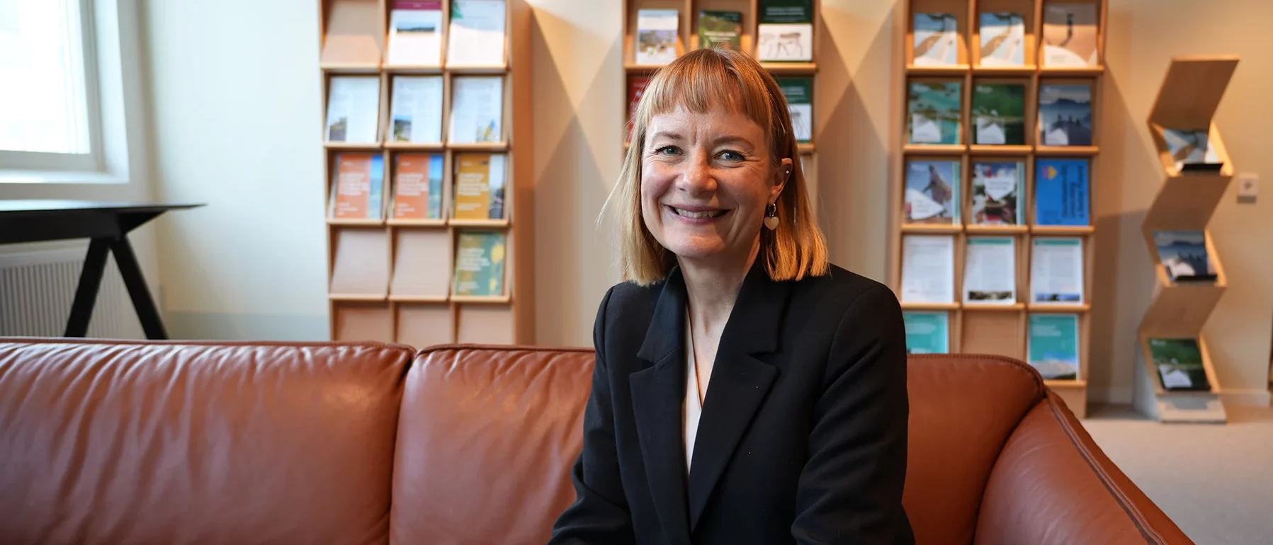 A woman in a black blazer sits smiling on a tan leather sofa, with display shelves of SEI publications behind her.