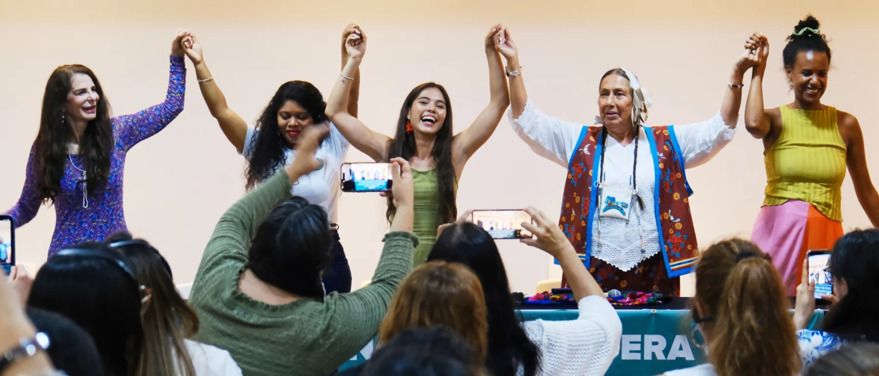 A row of smiling women behind a table on a stage stand and join lifted hands before an audience at the Santa Marta, Colombia, First Conference on Transitioning Away from Fossil Fuels.