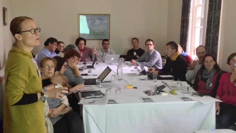 A woman in a yellow-green top stands at the head of a long table speaking to about a dozen people seated around it with laptops and notebooks.