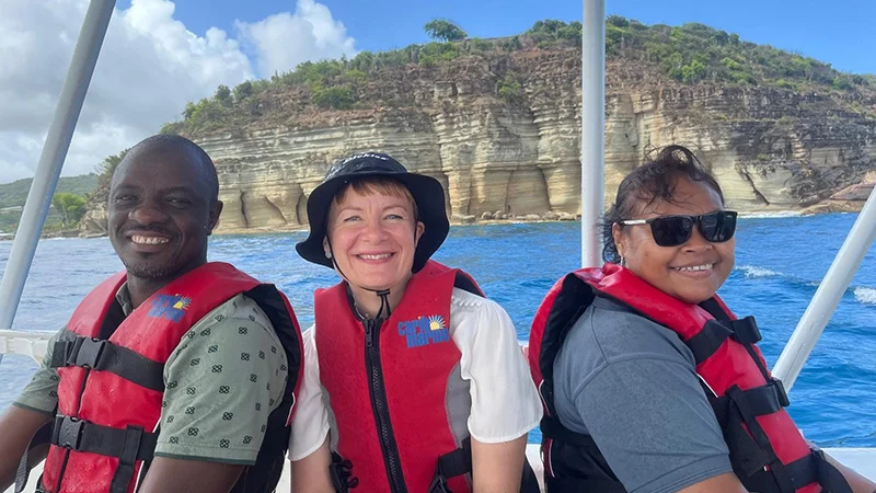 Three people in red life jackets sitting on a boat, smiling, with sandstone cliffs and blue sea behind them.