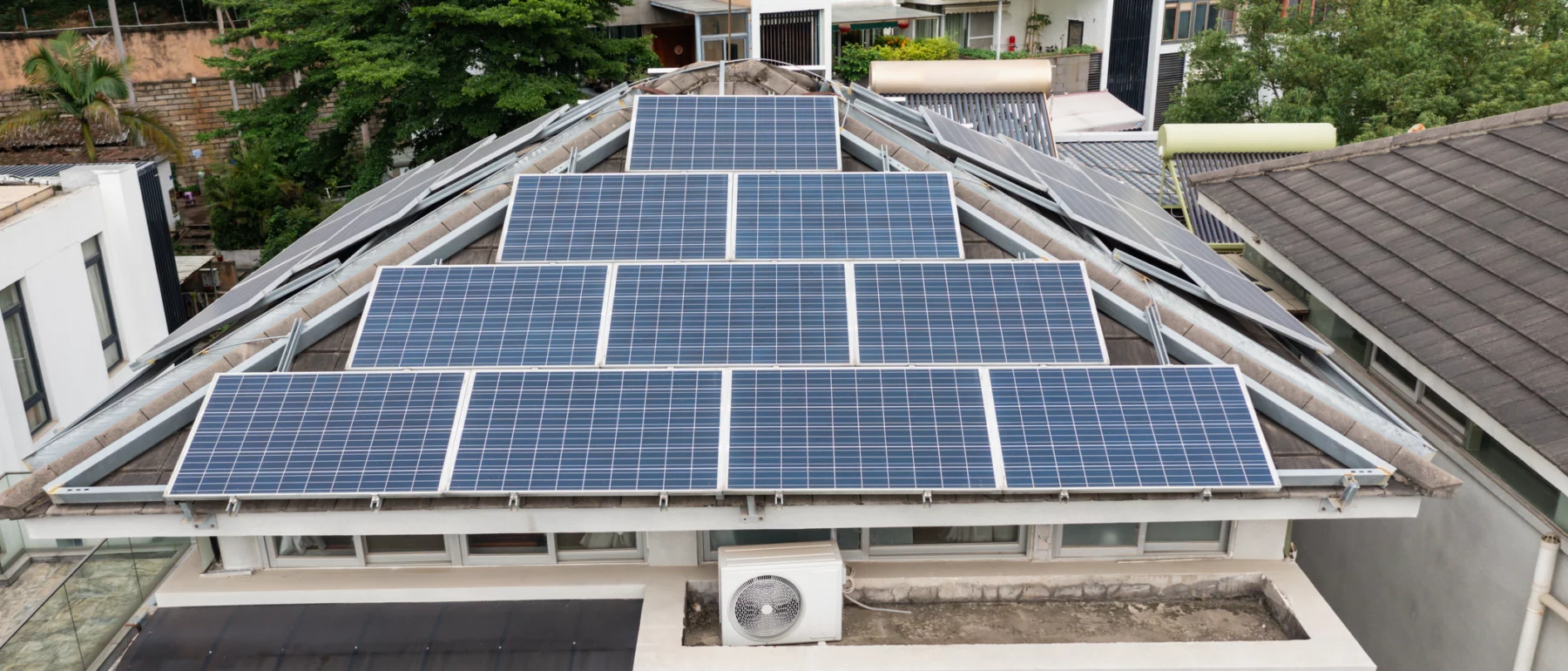 Solar panels mounted across a residential rooftop, with apartment buildings and trees in the background.