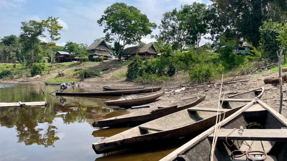 Wooden canoes line up in the foreground, beached below a collection of wooden houses among green trees.