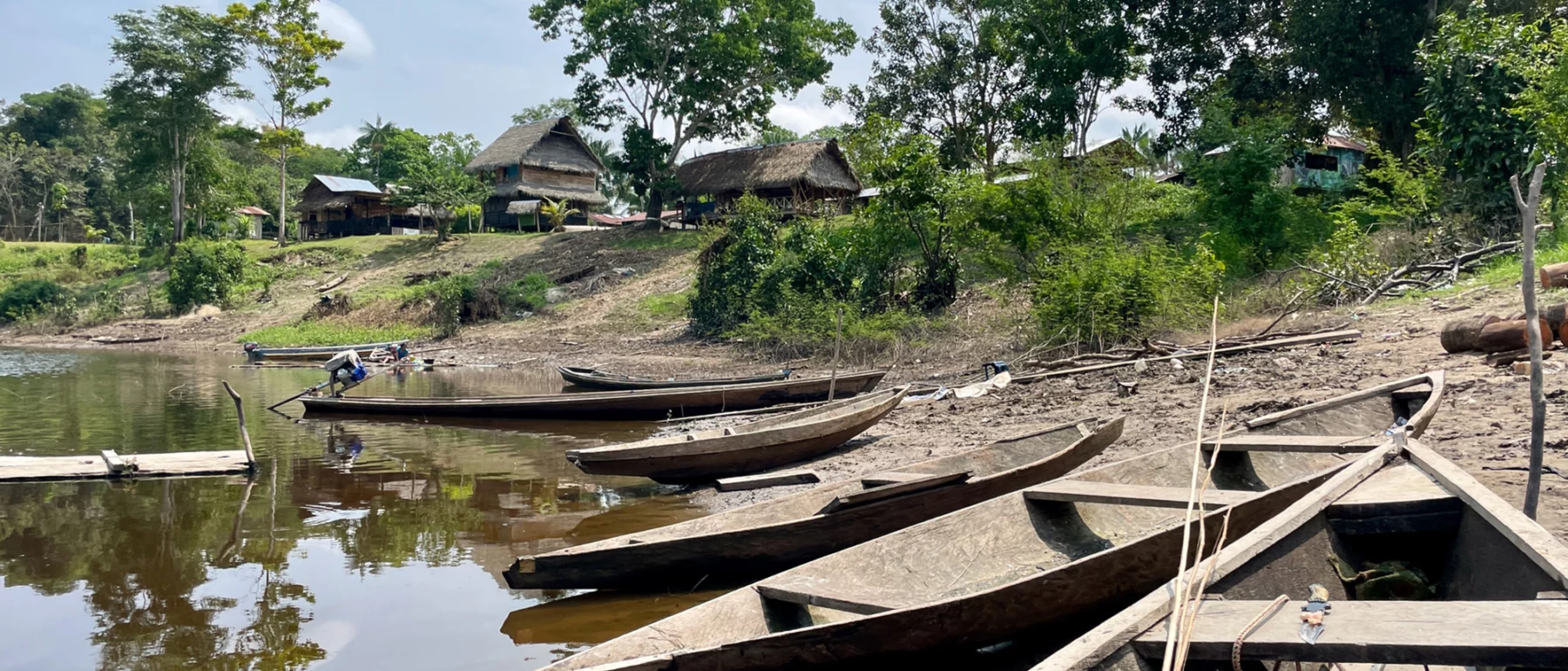 Wooden canoes line up in the foreground, beached below a collection of wooden houses among green trees.