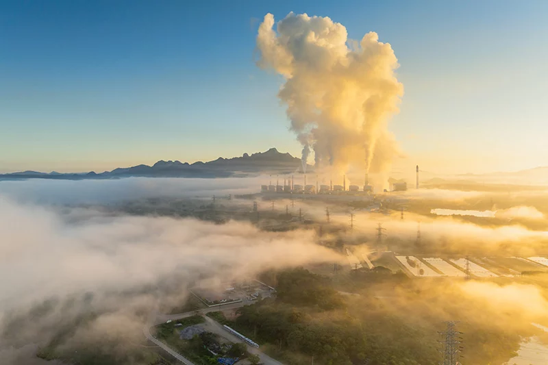 Aerial view of a coal-fired power plant with multiple chimneys releasing plumes of smoke at sunrise, surrounded by low-lying mist, forest and mountains in the distance.