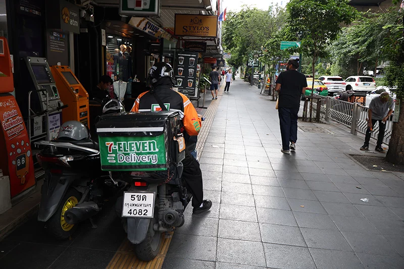 A delivery rider in an orange jacket parked on a Bangkok pavement beside a motorbike fitted with a 7-Eleven Delivery box, next to shopfronts and ATMs, with pedestrians walking along the street.