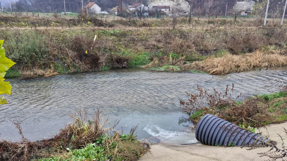 Water flows out of a drain into a stream in the foreground, which is flowing past a shrub-covered bank and a cluster of white farm buildings in the background on the other side of the stream.
