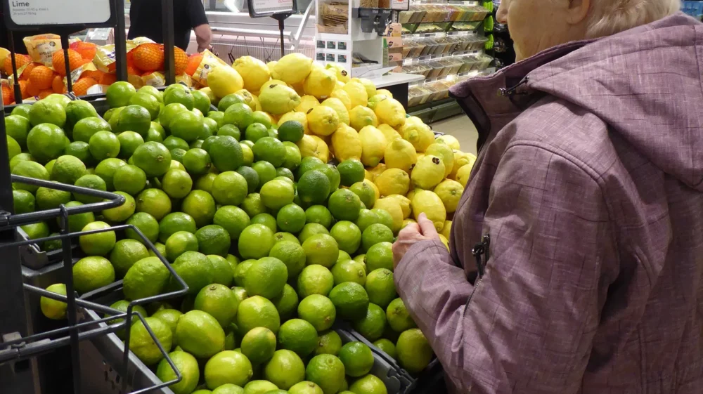 An older woman looks at bright green limes and yellow lemons stacked at a grocery store, with a sign in red and white in Swedish that says 4 for 10 SEK.