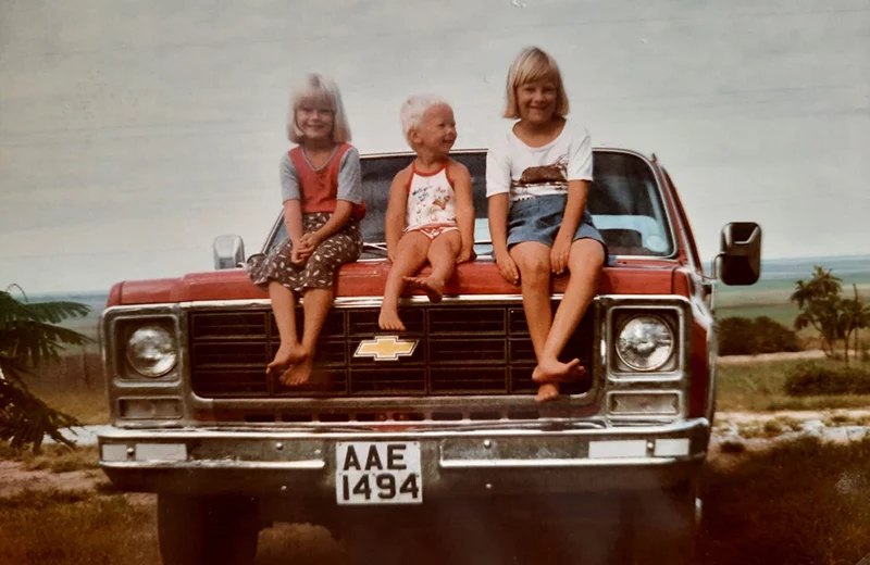 Three young children sitting barefoot on the bonnet of a red Chevrolet pickup truck, with open landscape behind.