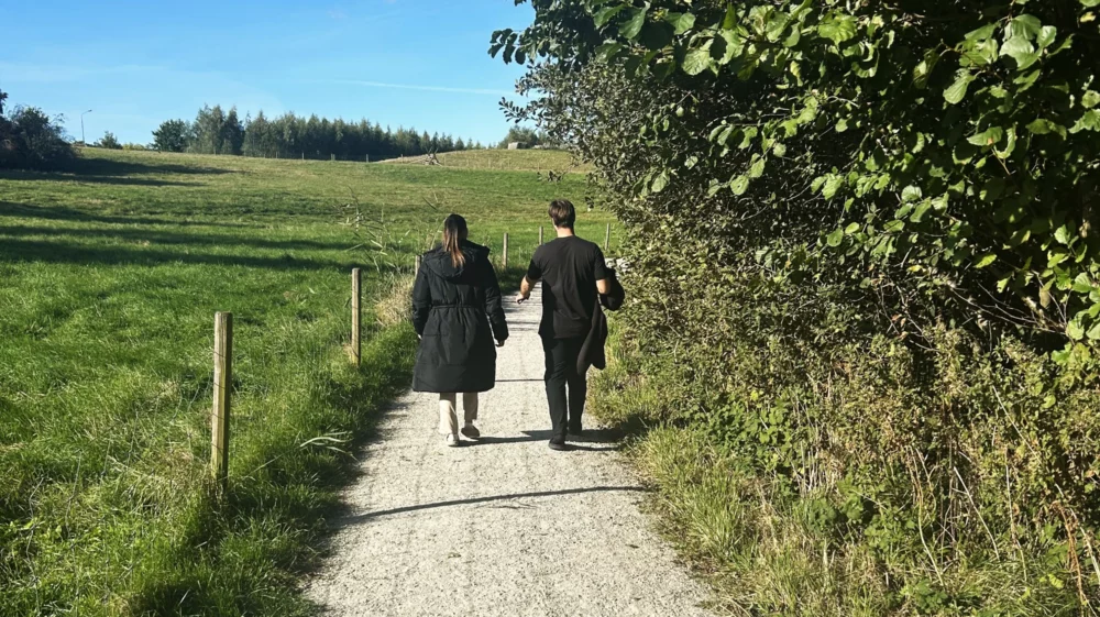 Two people dressed in black walk away along a gravel road. On the left is fenced grazing land for animals, and on the right side of the road are trees.