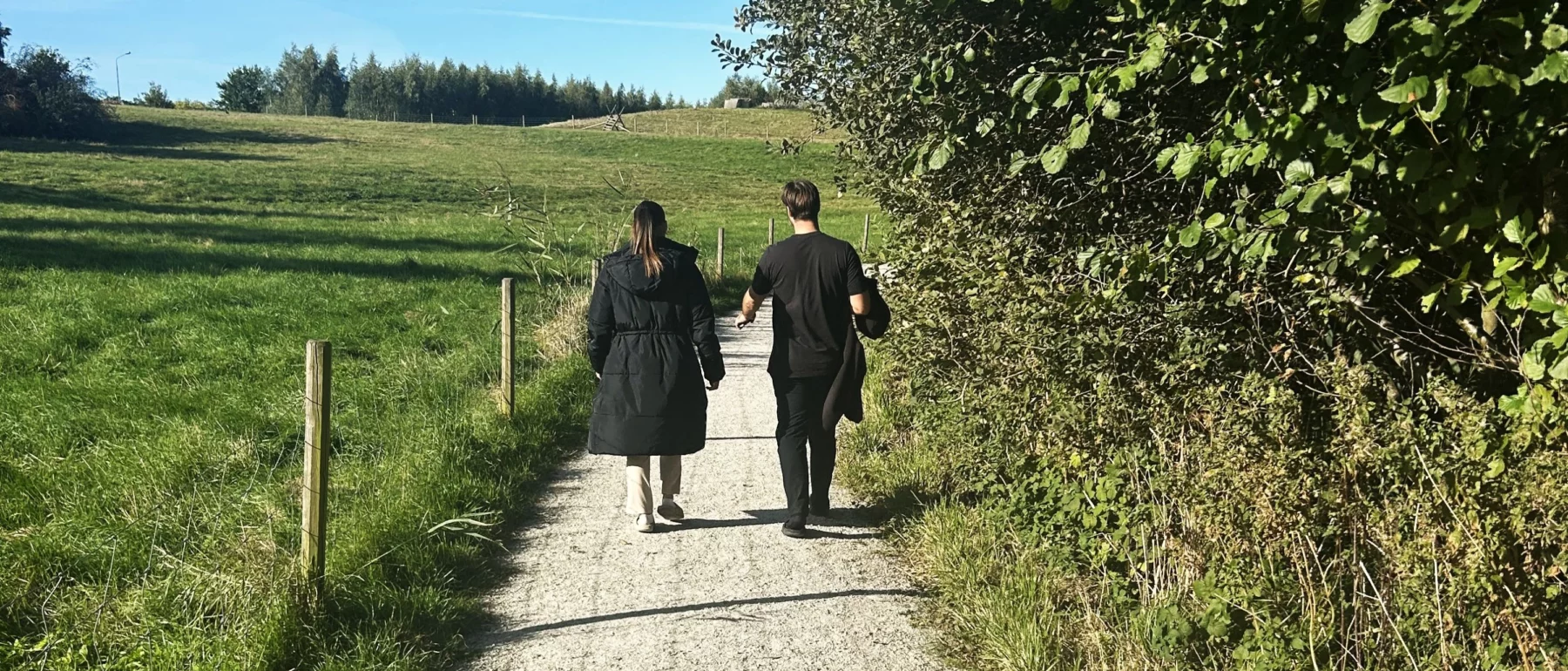 Two people dressed in black walk away along a gravel road. On the left is fenced grazing land for animals, and on the right side of the road are trees.