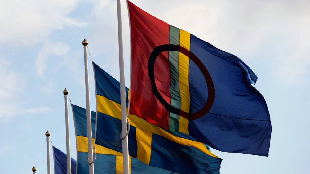 The red-green-yellow-blue striped flag with the black circle of the Sami flies in the foreground, with several blue and yellow Swedish flags behind.