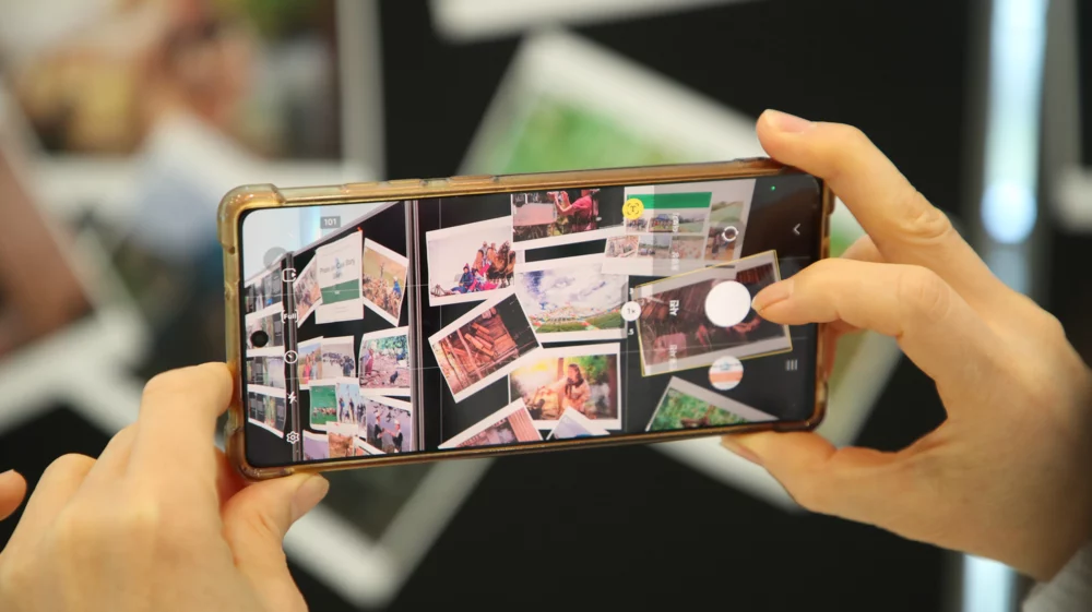 Hands holding a smartphone, photographing a scattered pile of printed photos on a table.