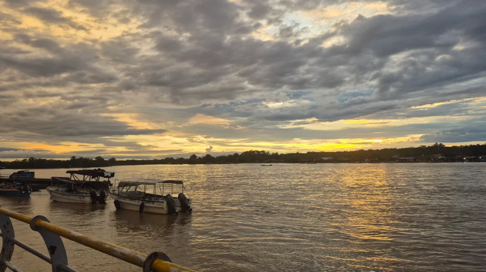 Blue grey clouds stretch to the yellow glow of the setting sun at the horizon, reflected in the river below, with several boats in the foreground and a quay, and small lights indicating houses and boats at the dark treelined bank on the far side.