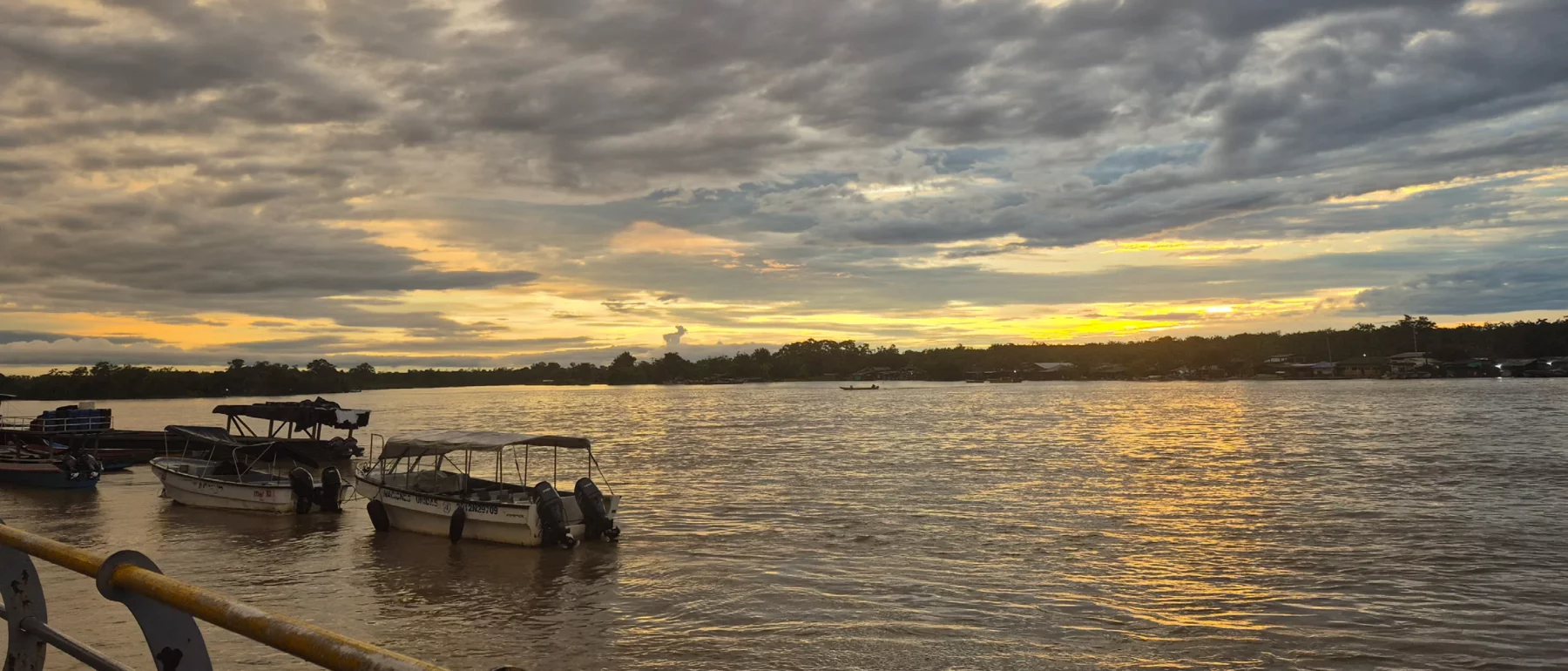 Blue grey clouds stretch to the yellow glow of the setting sun at the horizon, reflected in the river below, with several boats in the foreground and a quay, and small lights indicating houses and boats at the dark treelined bank on the far side.