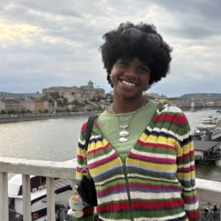 Chloe Daniel stands in front of a waterfront under an overcast sky, smiling, wearing a brightly colored horizontal striped zip-up sweater over a jade green shirt with a silver necklace.