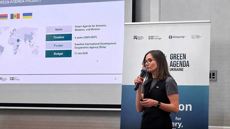 A woman wearing glasses speaks into a microphone beside a Green Agenda Ukraine banner, with a project overview slide projected behind her.