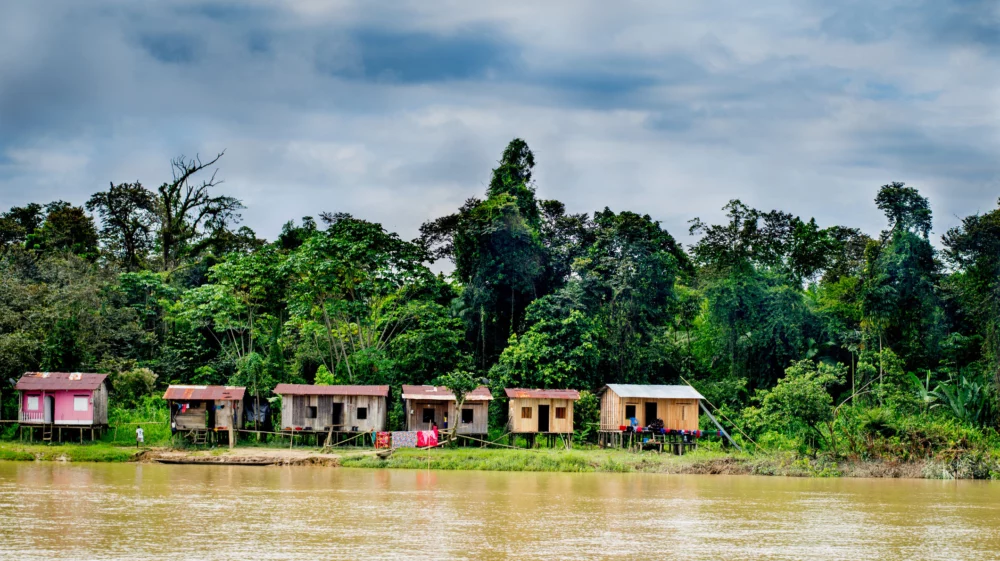 Landscape shot across a broad brown river. On the other side are small wooden houses on stilts. Colourful laundry hangs on washing lines in front of them. Behind them are tall, lush green trees underneath a blue sky dappled with light cloud.