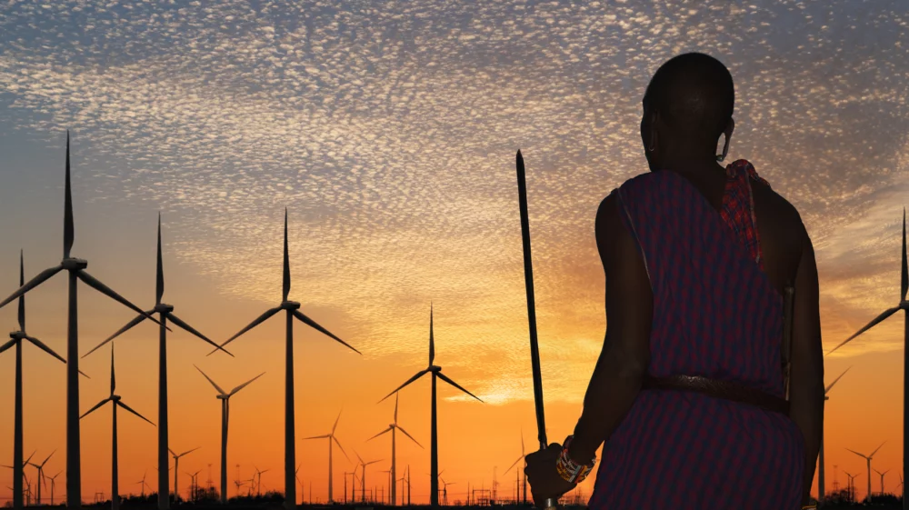 Rear view of a Masai warrior in Nairobi, Kenya wearing traditional clothes watching silhouetted wind turbines at sunset beneath a dark blue and bright orange, lightly clouded sky