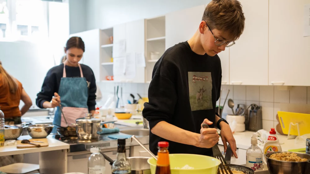 Avar kooliköök, mille tööpinnad on kaetud erinevate kausside, pottide, kastmepudelite ja muude toiduvalmistamise vahenditega. Esiplaanil valmistab noormees midagi pannil. Tagaplaanil valmistab neiu midagi pannil.