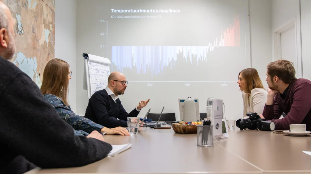 People in discussion around a meeting table, with a chart of global temperature change projected on the wall behind them.