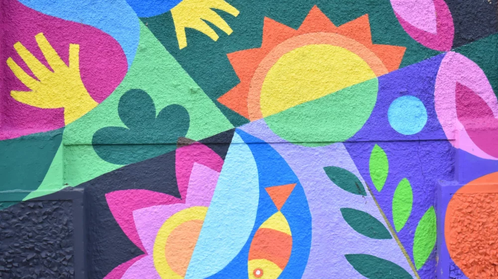 Brightly coloured artwork painted on wall featuring simplistic images of plants, a fish, the sun and a human hand.