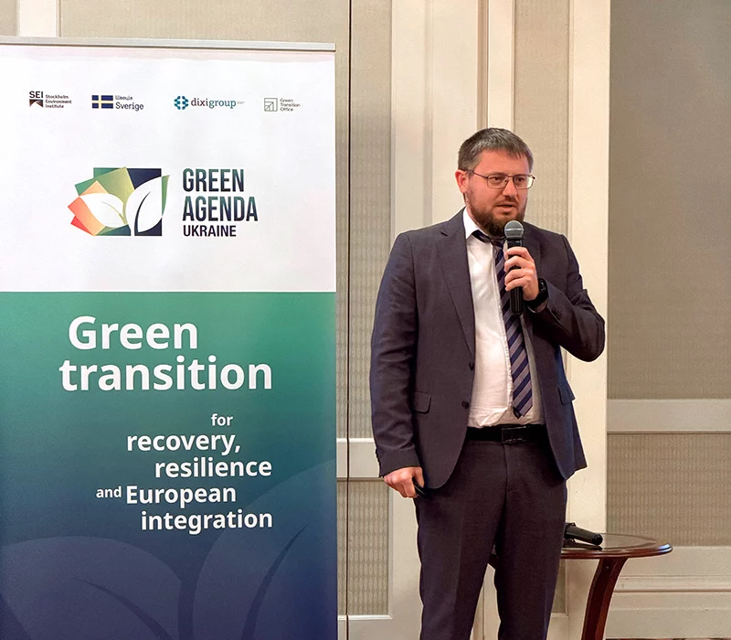 A man in a dark suit and striped tie speaks into a microphone next to a Green Agenda Ukraine banner.