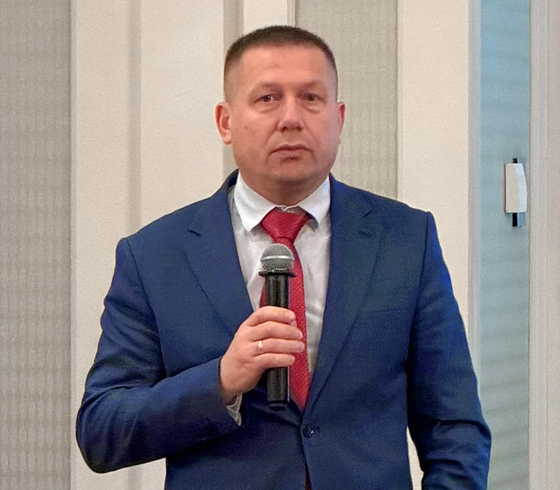 A man in a blue suit and red tie holds a microphone as he addresses the room.