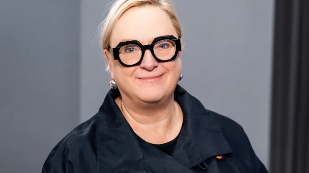 A headshot of a woman with short blonde hair and dark thick glasses smiling at the camera.