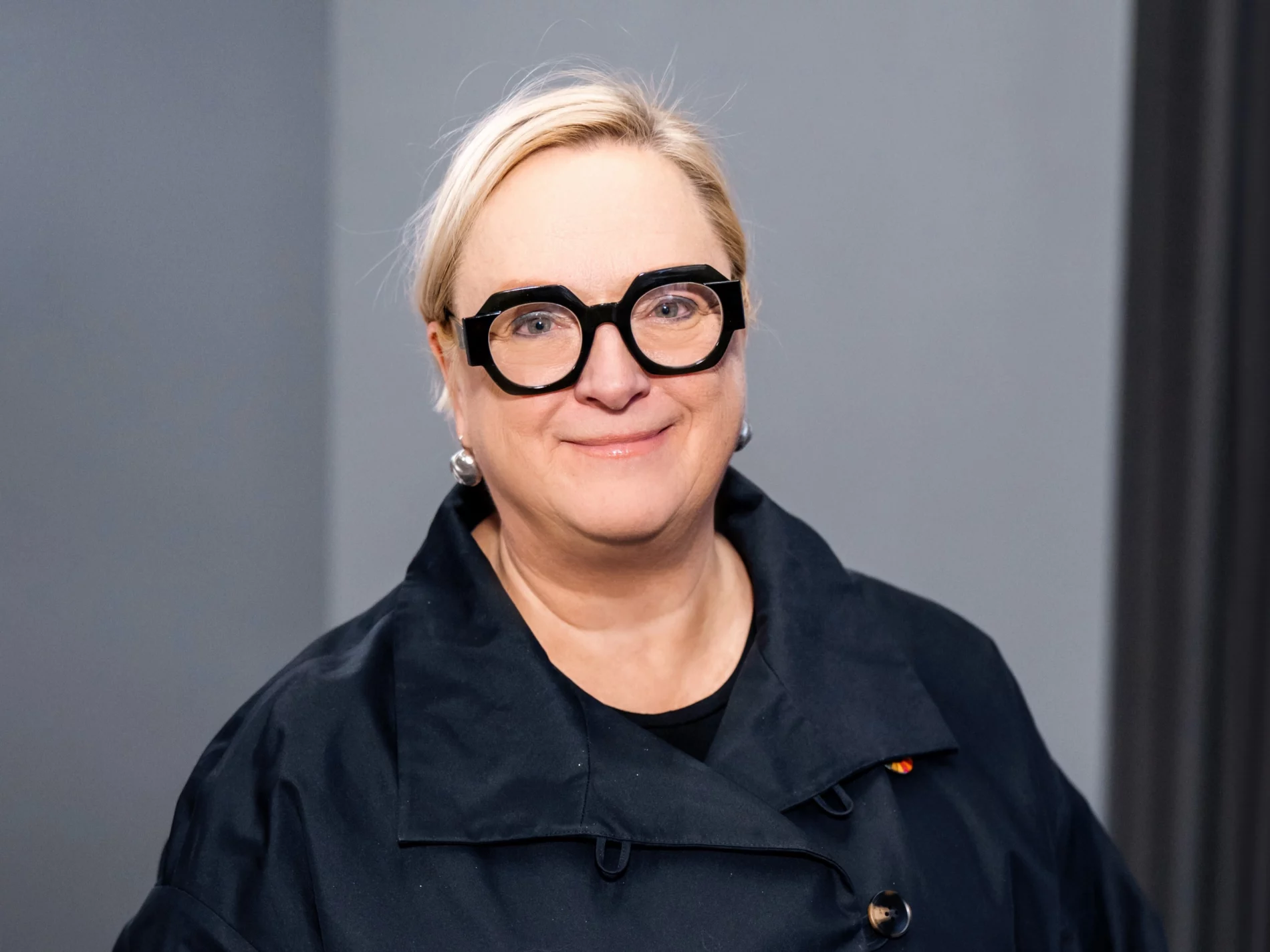 A headshot of a woman with short blonde hair and dark thick glasses smiling at the camera.