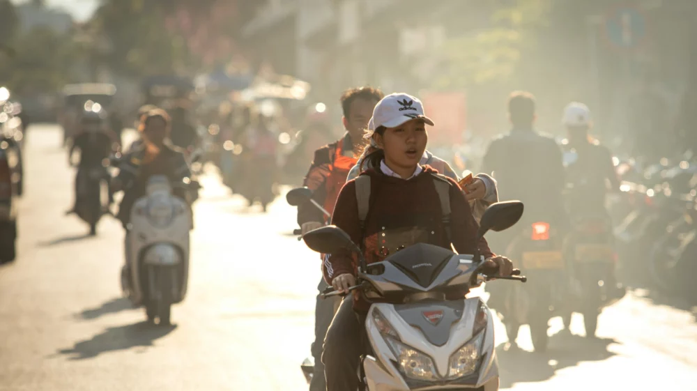 A young woman rides a motorbike through a busy street at golden hour, with other riders behind her.