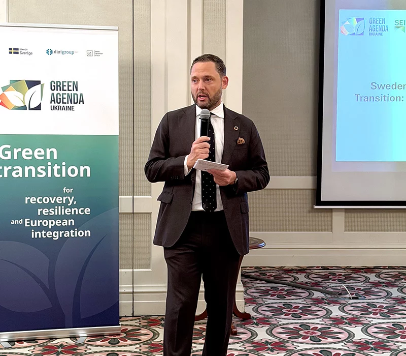 A man in a dark suit holds a microphone and notes while speaking beside a Green Agenda Ukraine banner.