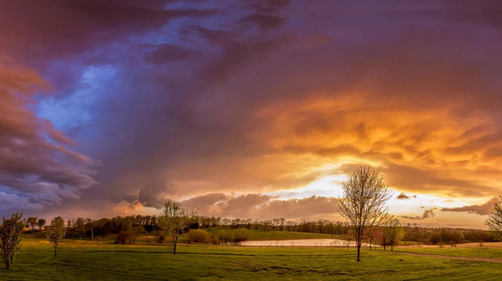 Panorama of green grass and deciduous trees beneath a colorful cloudy sky that transitions from blue on the left to orange on the right, where the sun peeks through.