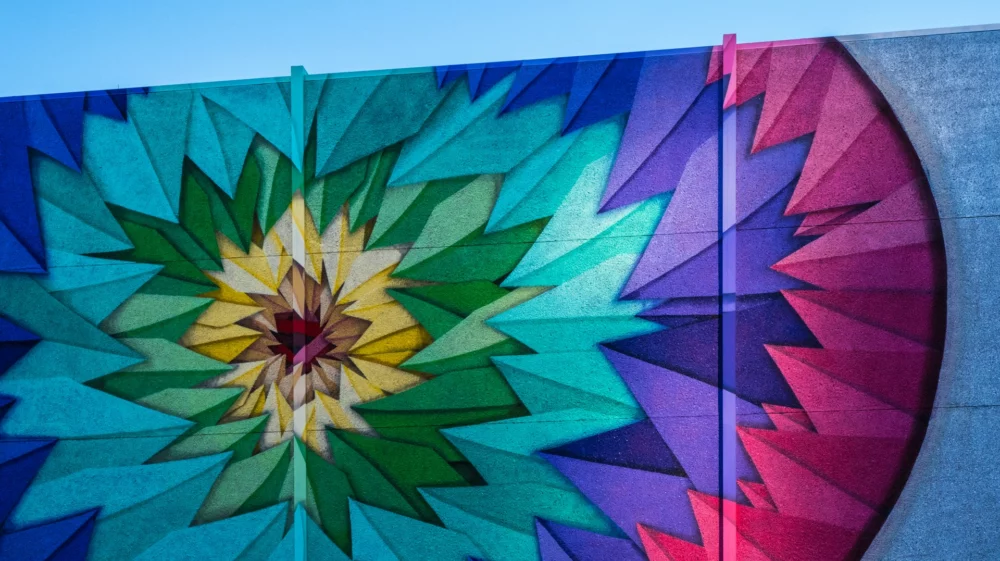 Colourful mural on a wall under a blue sky: jagged, concentric oval rings move inwards: pink, purple, light blue, green, yellow and red in the middle. The effect almost looks like a flower.