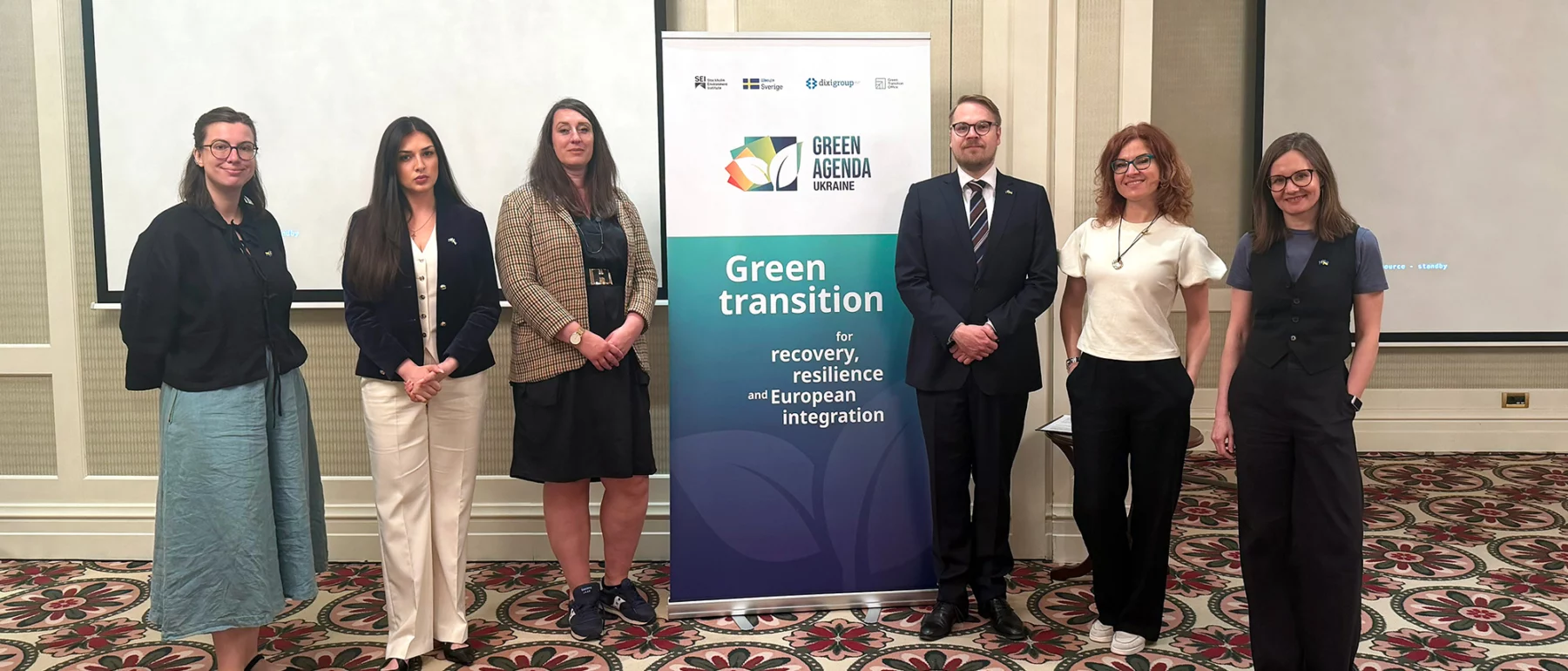 Six people stand on either side of a Green Agenda Ukraine banner in a conference room with a patterned carpet.