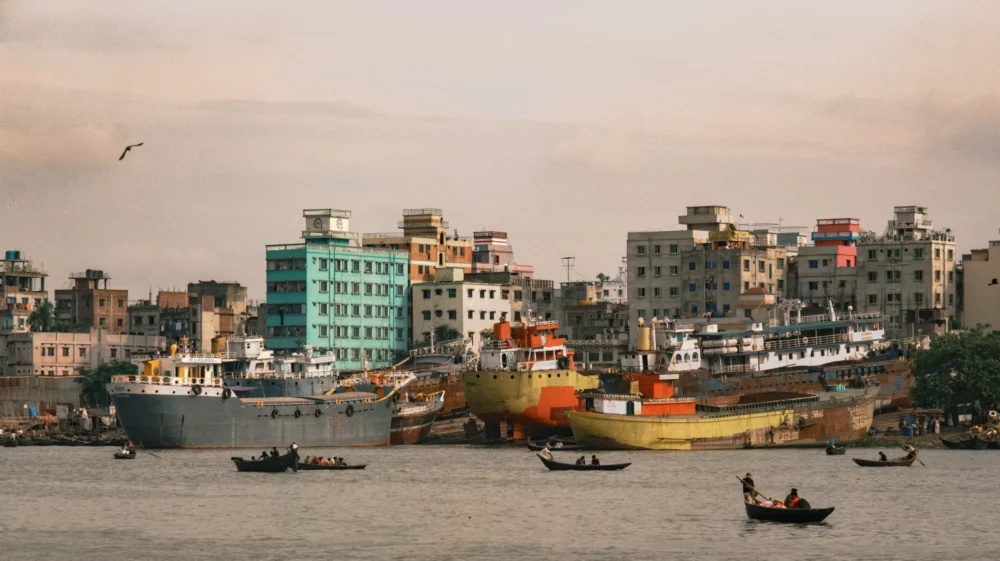 On the far bank of a river, white, brown and tan buildings are punctuated by a aquamarine-green one, with grey, white and orange ships on the water's edge below, small boats, trees and people.