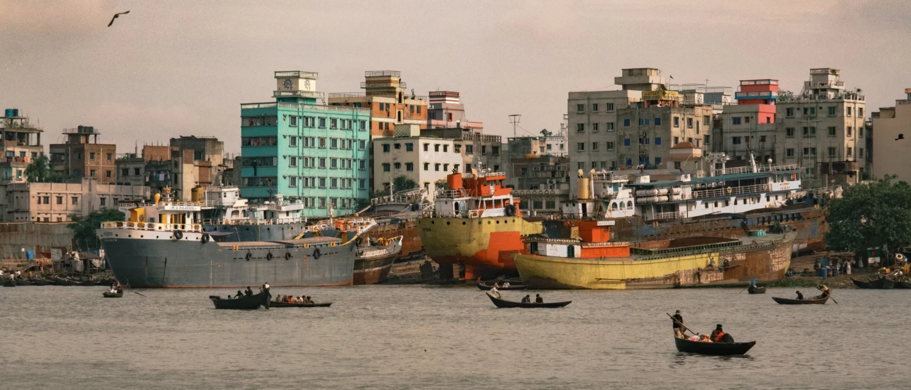 On the far bank of a river, white, brown and tan buildings are punctuated by a aquamarine-green one, with grey, white and orange ships on the water's edge below, small boats, trees and people.