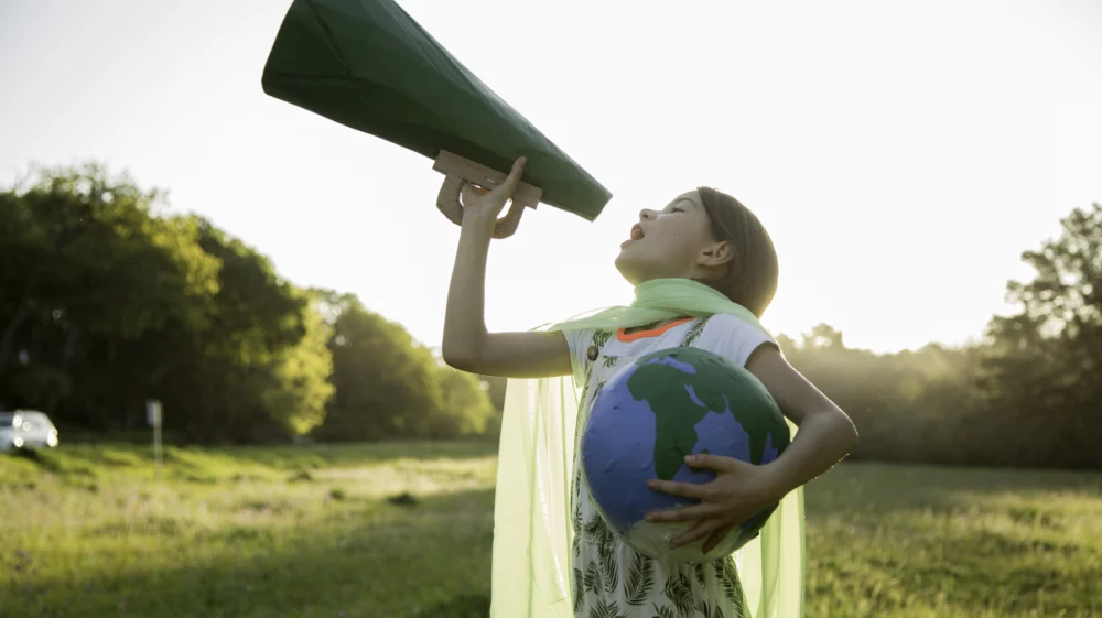 Girl with brown hair about 8 to 10 years old wearing a scarf fashioned into a cape and holding a globe in her left arm while hoisting a green megaphone with her right, appearing to shout into it