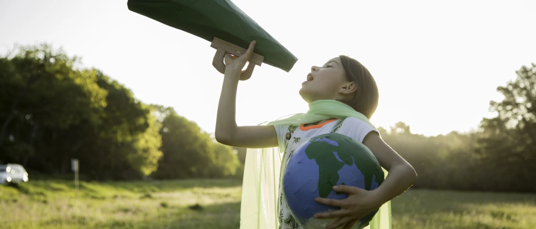 Girl with brown hair about 8 to 10 years old wearing a scarf fashioned into a cape and holding a globe in her left arm while hoisting a green megaphone with her right, appearing to shout into it