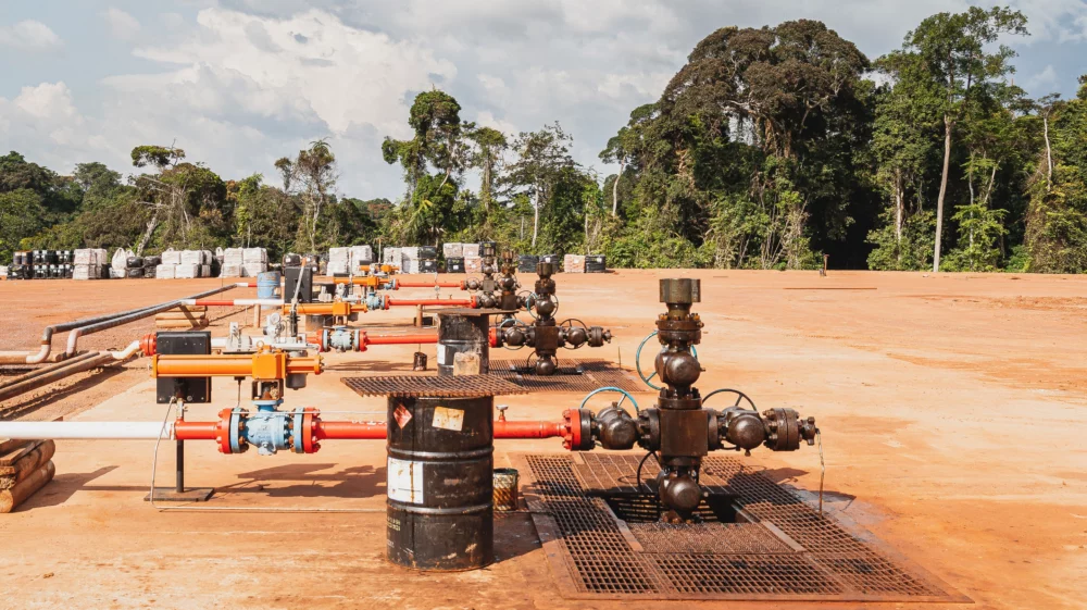 A network of multi-coloured gas pipes and black barrels in an open dirt clearing, with trees in the background.