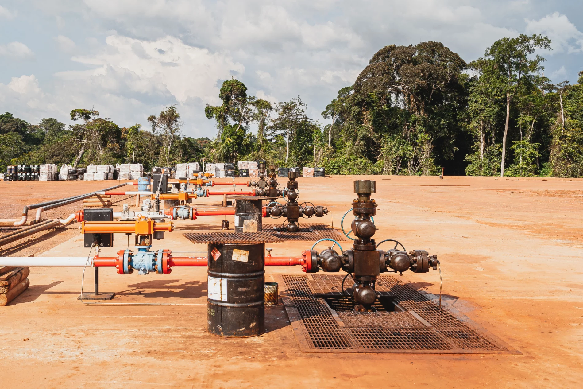 A network of multi-coloured gas pipes and black barrels in an open dirt clearing, with trees in the background.