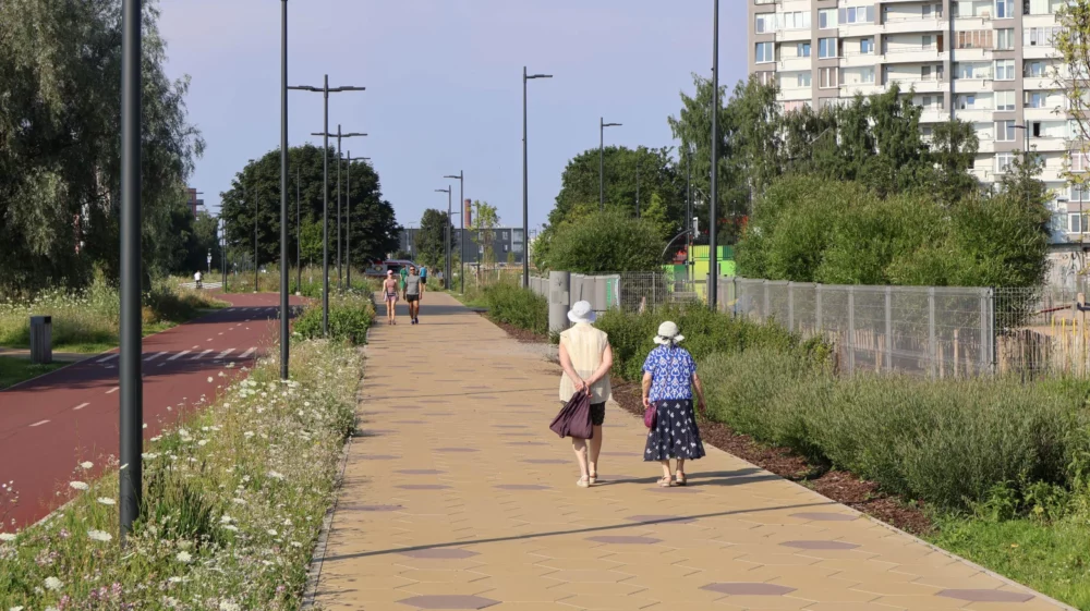 A paved pedestrian path runs alongside a separate red-surfaced bike lane, bordered by wildflower meadows and tall grasses. Two older women in sun hats and floral clothing walk away from the camera on the path, with two joggers approaching in the distance. Rows of streetlights line the path, with a tall apartment block visible on the right and green trees along the left.