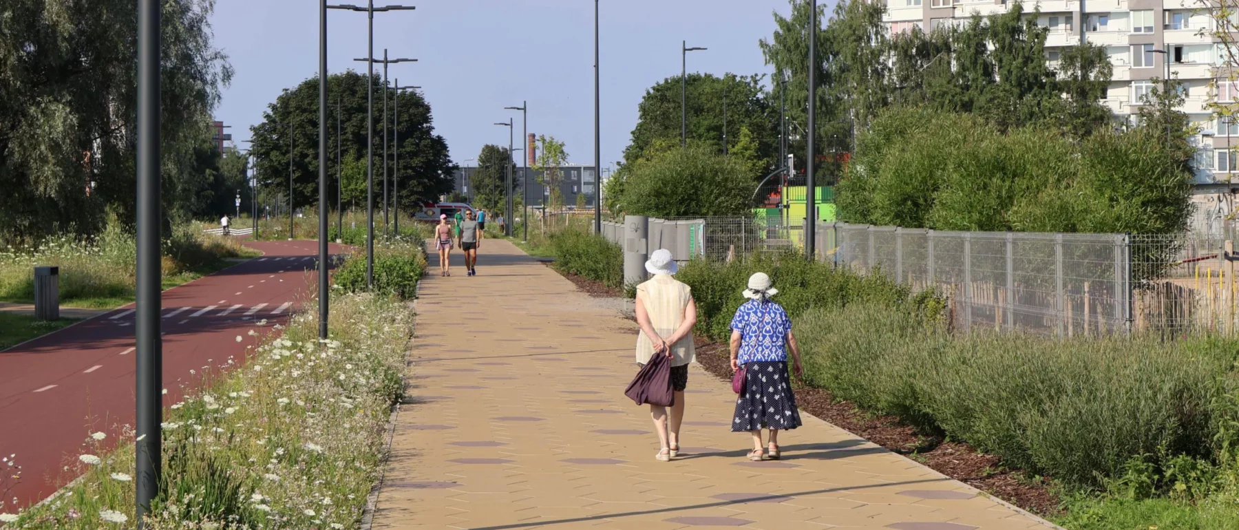A paved pedestrian path runs alongside a separate red-surfaced bike lane, bordered by wildflower meadows and tall grasses. Two older women in sun hats and floral clothing walk away from the camera on the path, with two joggers approaching in the distance. Rows of streetlights line the path, with a tall apartment block visible on the right and green trees along the left.