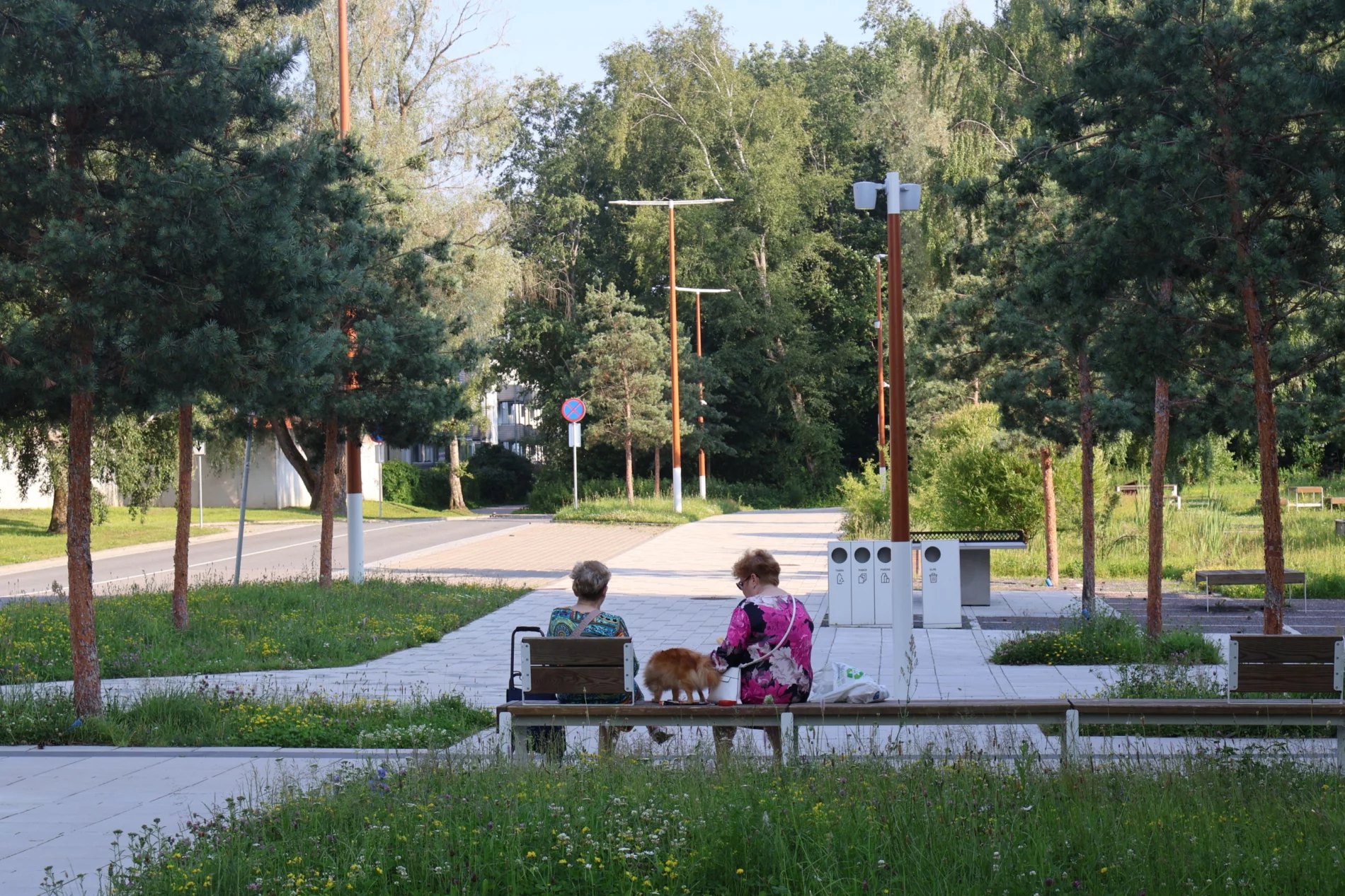 Kaks naist istuvad koos koeraga pargipingil rohelise puiestee ääres, ümbritsetud puude ja lilledega.