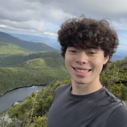 Selfie showing Lincoln smiling, with curly dark hair wearing a dark T-shirt standing at a viewpoint over rolling green hills, a cloudy blue sky and a body of water.