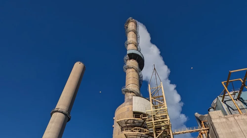 Two industrial chimneys reach up into a blue sky, with a plume streaming out of the center-right stack, looming over industrial buildings with bright yellow doors and ladders.
