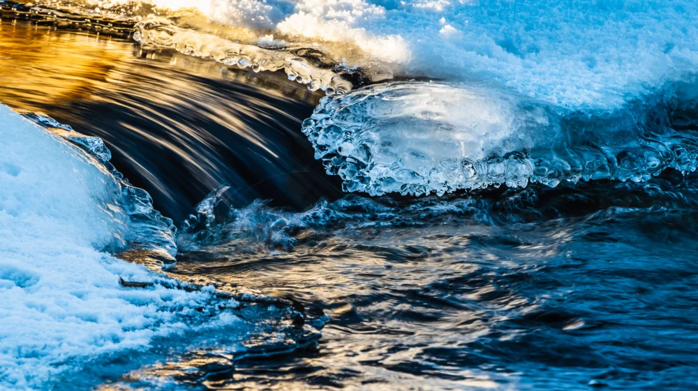 Dark flowing water creates white foam as it crashes into calm water, moving left to right, with ice on either side.