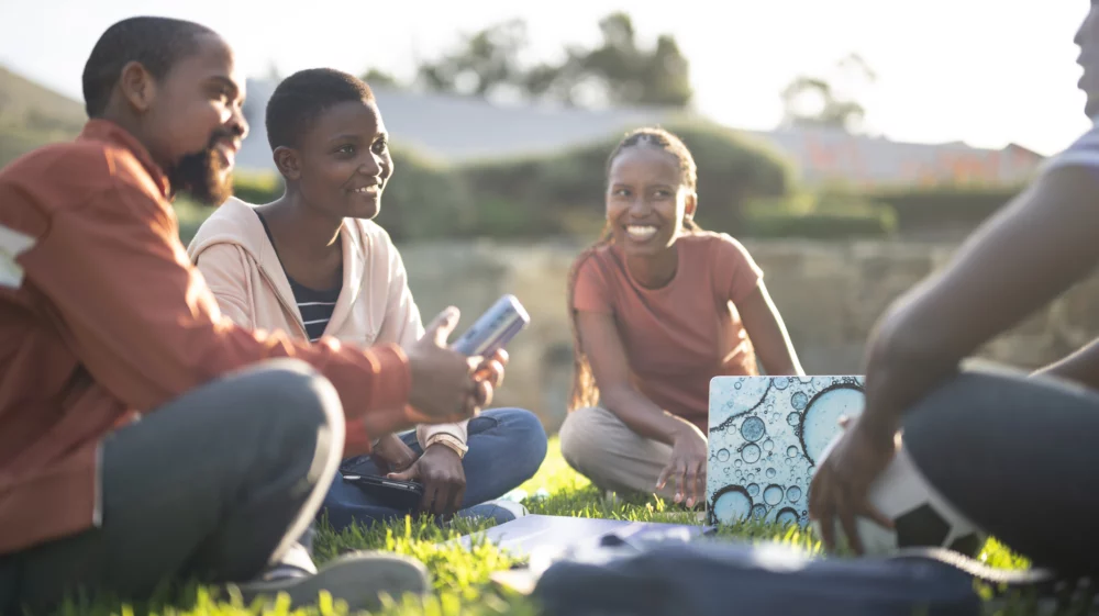 A group of university-age students sit on a lawn with smart phones and a lap top computer, smiling